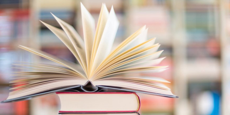 A Book Opened Which Is Placed On Another Book In A Blurred Library background.