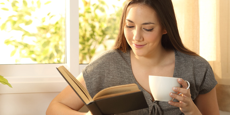 A Smiling Young Lady Reads A Book And Have A Cup of Coffee In Her Hand.